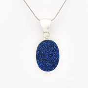 A silver pendant necklace featuring an oval-shaped, glittery royal blue stone, suspended from a delicate silver chain against a plain white background.