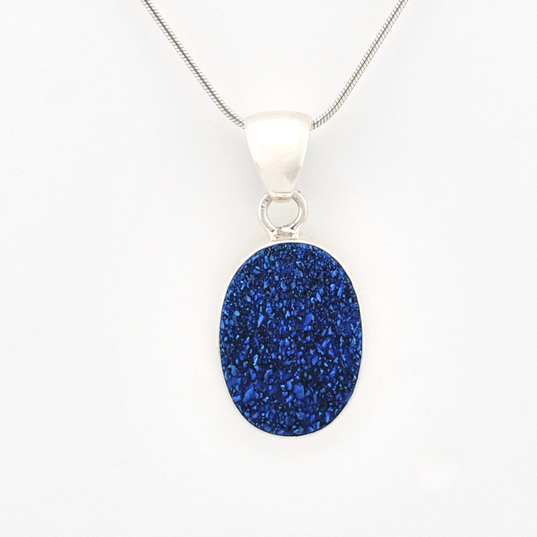 A silver pendant necklace featuring an oval-shaped, glittery royal blue stone, suspended from a delicate silver chain against a plain white background.