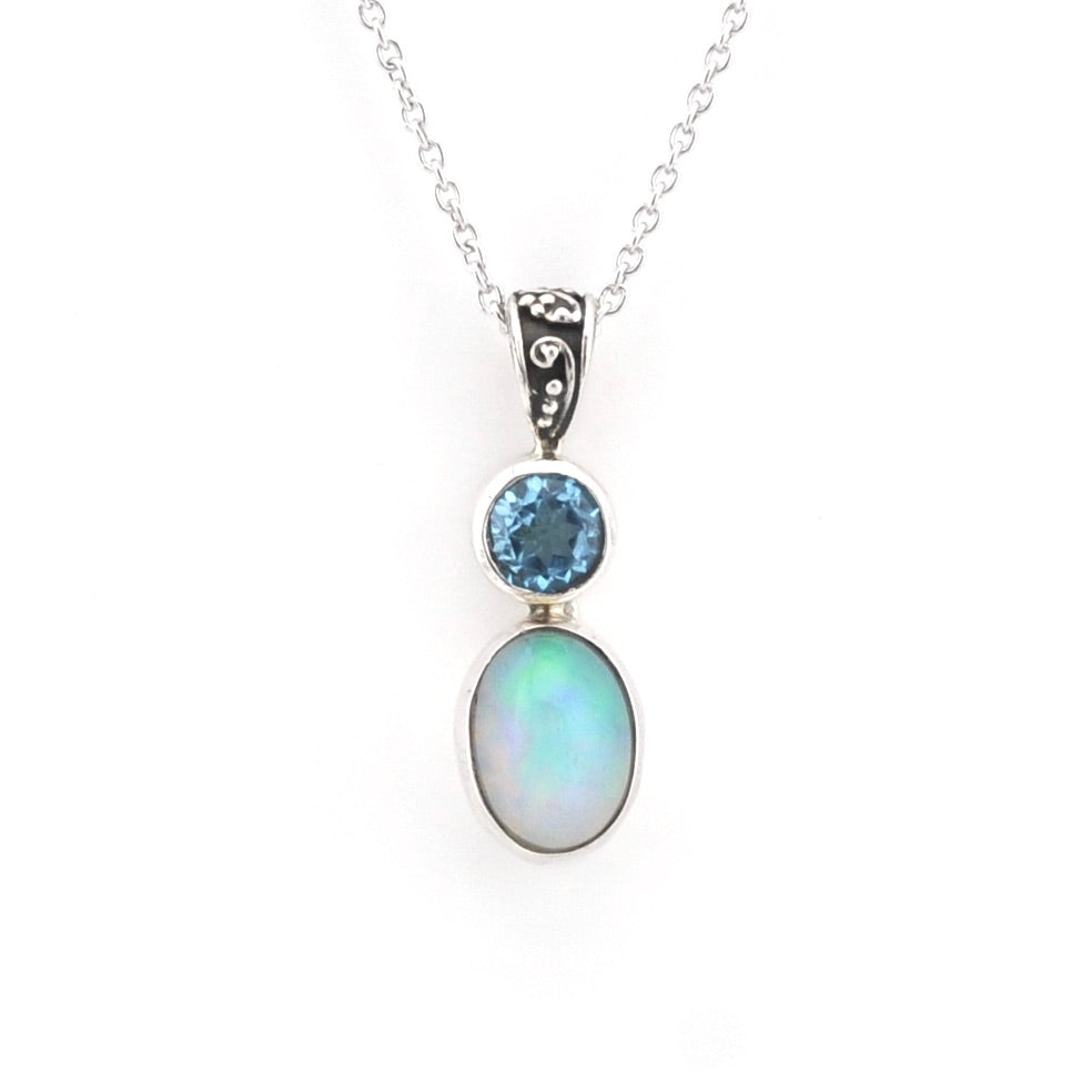 A silver pendant necklace featuring a round blue topaz stone and an oval iridescent opal gemstone in a silver bezel setting, suspended from a delicate silver chain against a white background.