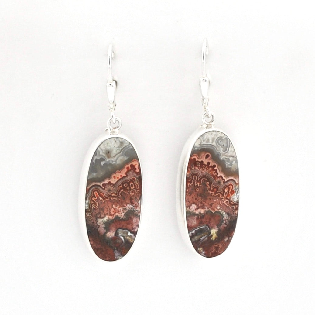 A pair of silver drop earrings featuring oval-shaped cabochon gemstones with a natural agate or jasper pattern in shades of brown, gray, and black, suspended from silver fishhook wires against a plain white background.