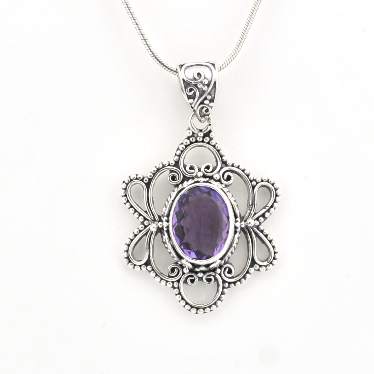A silver filigree pendant necklace featuring an oval purple amethyst gemstone in the center, suspended from a delicate silver snake chain against a plain white background.