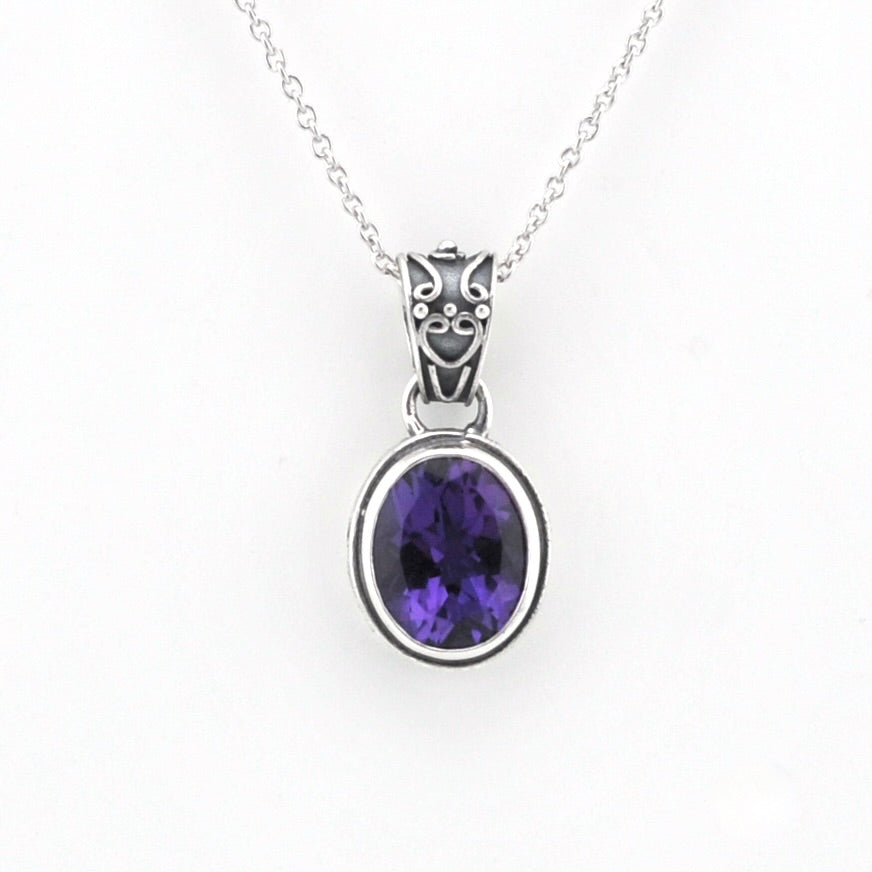 A silver pendant necklace featuring an oval-cut purple gemstone in a silver bezel setting, suspended from a silver chain with an ornate silver bail.