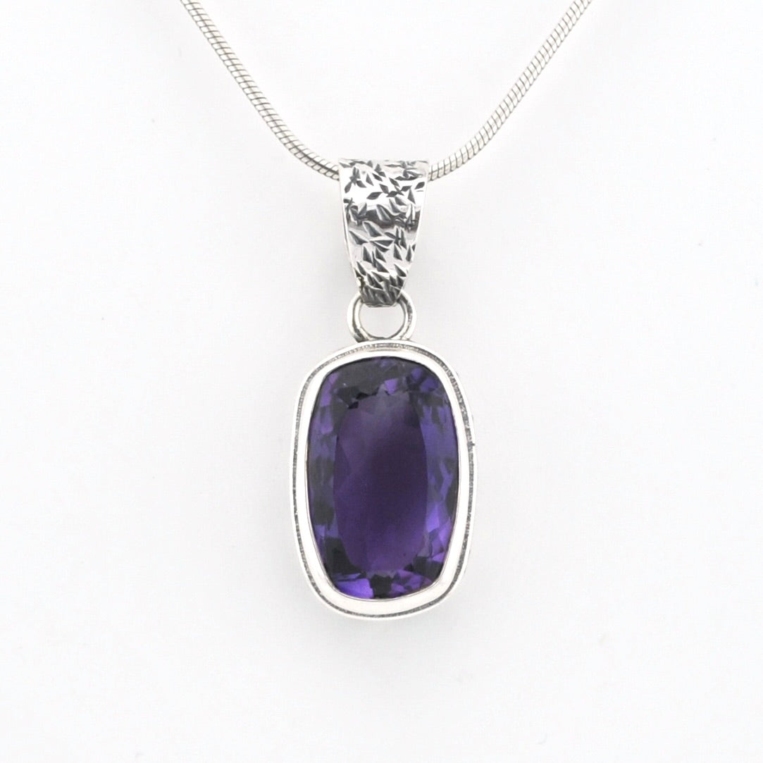 A silver pendant necklace featuring a bezel-set oval purple gemstone on a delicate silver chain, shown against a plain white background.
