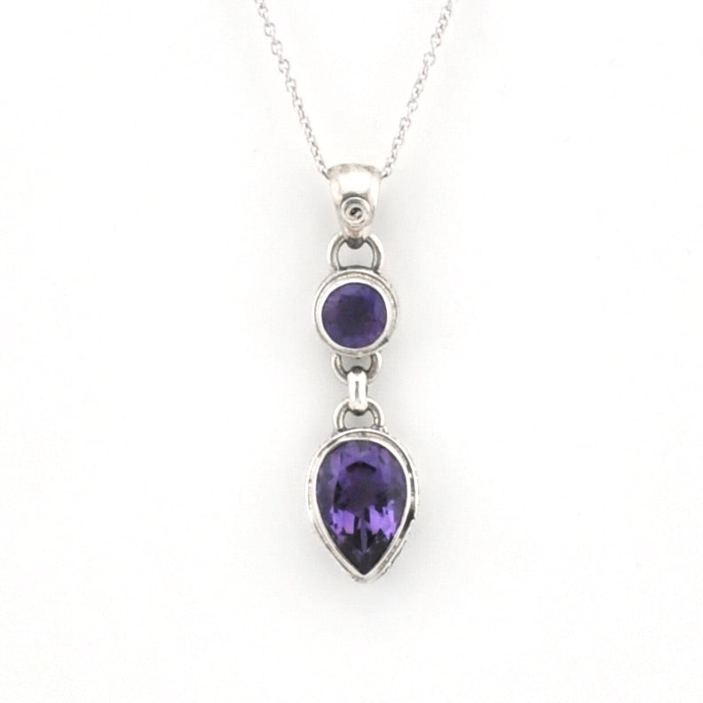 A silver pendant necklace featuring a teardrop-shaped purple gemstone and a smaller oval purple gemstone set in a silver bezel, suspended from a delicate silver chain against a white background.