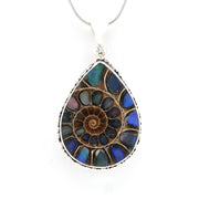 A teardrop-shaped pendant necklace featuring a spiral ammonite fossil design in iridescent blue, green, and brown opal-like stones, set in a silver-toned bezel with a silver snake chain visible at the top, on a plain white background.