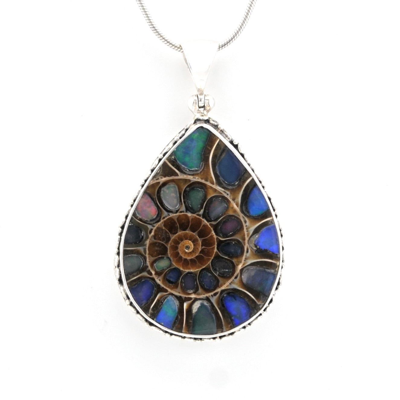 A teardrop-shaped pendant necklace featuring a spiral ammonite fossil design in iridescent blue, green, and brown opal-like stones, set in a silver-toned bezel with a silver snake chain visible at the top, on a plain white background.
