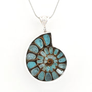 A silver pendant necklace featuring a spiral ammonite fossil inlay with turquoise blue, brown, and white natural stone patterns, suspended on a silver chain against a white background.