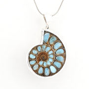 A silver pendant necklace featuring a polished ammonite shell inlay with light blue and brown natural stone segments, suspended from a silver chain against a plain white background.