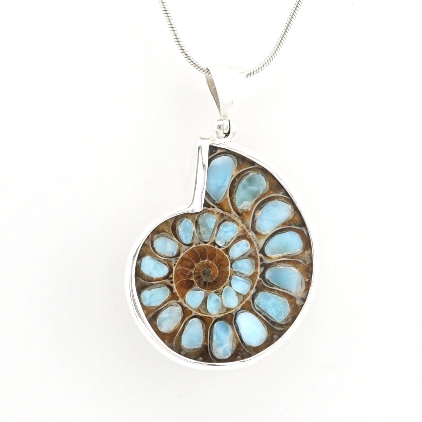 A silver pendant necklace featuring a polished ammonite shell inlay with light blue and brown natural stone segments, suspended from a silver chain against a plain white background.