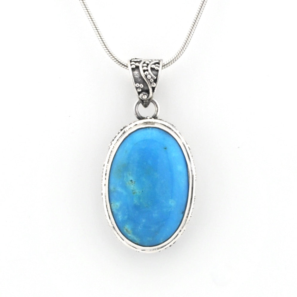 An oval pendant necklace featuring a vibrant blue cabochon gemstone in a silver bezel setting, suspended from a silver snake chain against a plain white background.