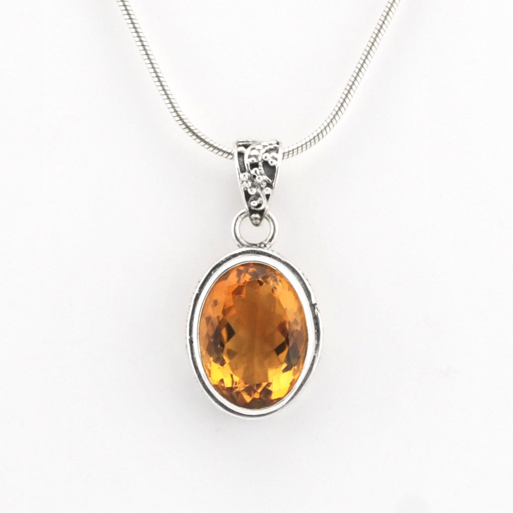 A silver pendant necklace featuring an oval-cut orange-yellow gemstone in a silver bezel setting, suspended from a silver snake chain against a plain white background.
