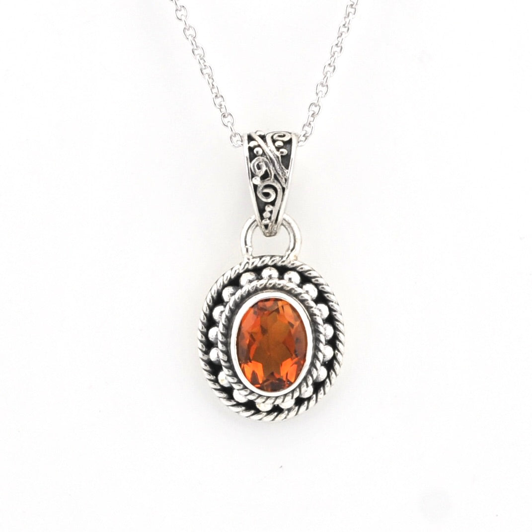 A silver pendant necklace featuring a oval-cut orange gemstone surrounded by a textured, oxidized silver border and a decorative filigree bail, suspended from a silver chain on a white background.