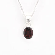 A silver pendant necklace featuring an oval-cut dark red garnet gemstone in a silver bezel setting, suspended from a delicate silver chain against a plain white background.