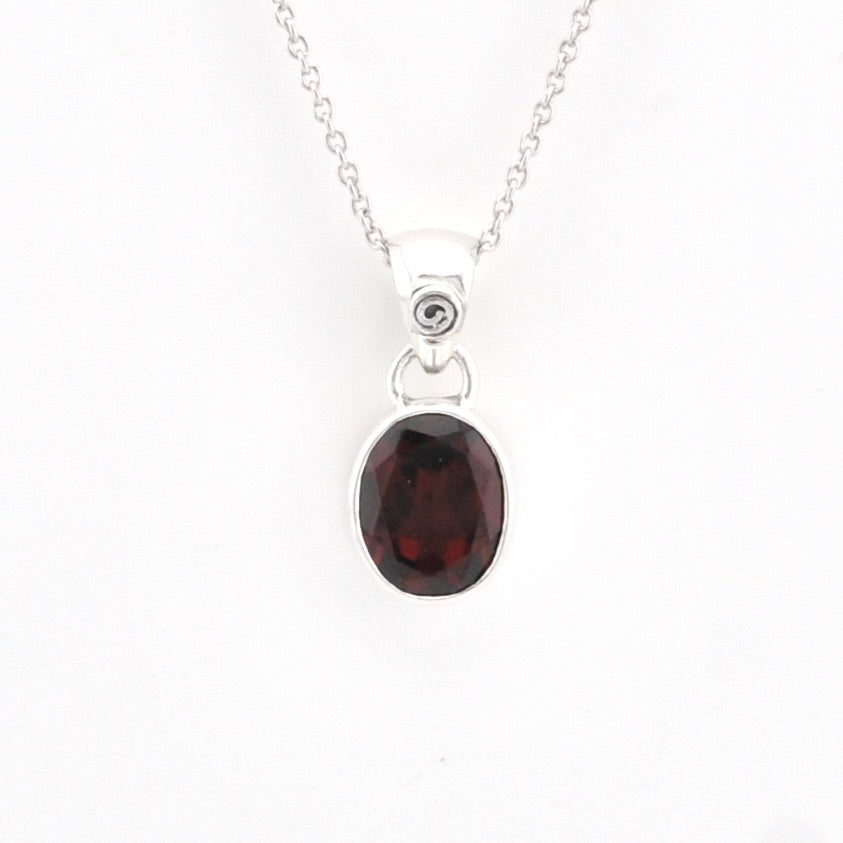 A silver pendant necklace featuring an oval-cut dark red garnet gemstone in a silver bezel setting, suspended from a delicate silver chain against a plain white background.