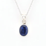 A silver pendant necklace featuring an oval-cut deep blue sapphire gemstone in a polished silver bezel setting, suspended from a delicate silver chain against a plain white background.