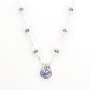 A delicate silver chain necklace featuring alternating white pearls and faceted purple beads, with a central teardrop-shaped pendant showcasing a blue and purple floral pattern on a white background.