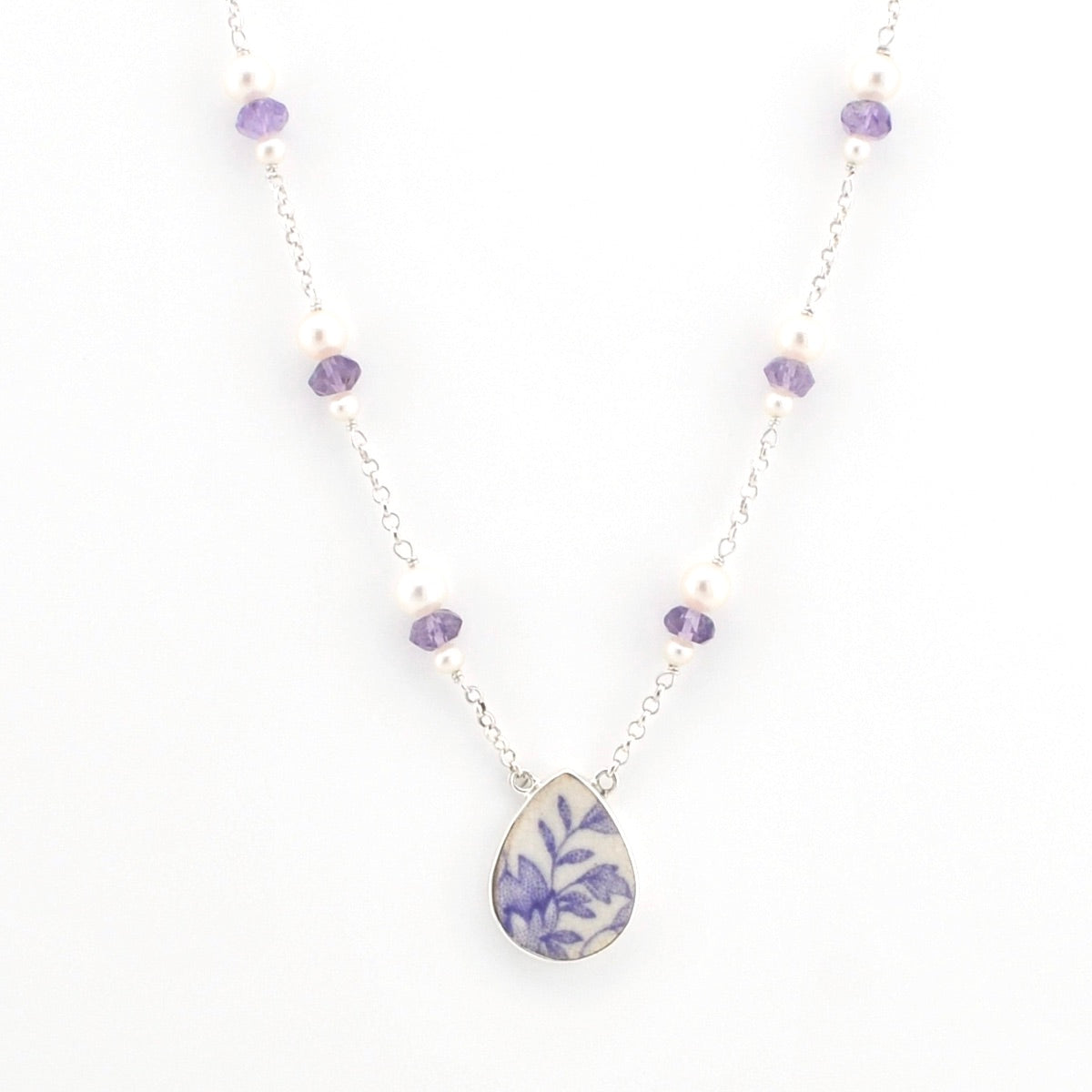 A delicate silver chain necklace featuring alternating white pearls and faceted purple beads, with a central teardrop-shaped pendant showcasing a blue and purple floral pattern on a white background.
