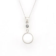 A silver pendant necklace featuring a large, smooth circular disc and a small round bezel-set dark stone, suspended from a delicate silver chain against a plain white background.