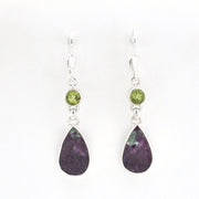 A pair of silver dangle earrings featuring a small round green gemstone and a larger teardrop-shaped dark purple stone, shown against a plain white background.