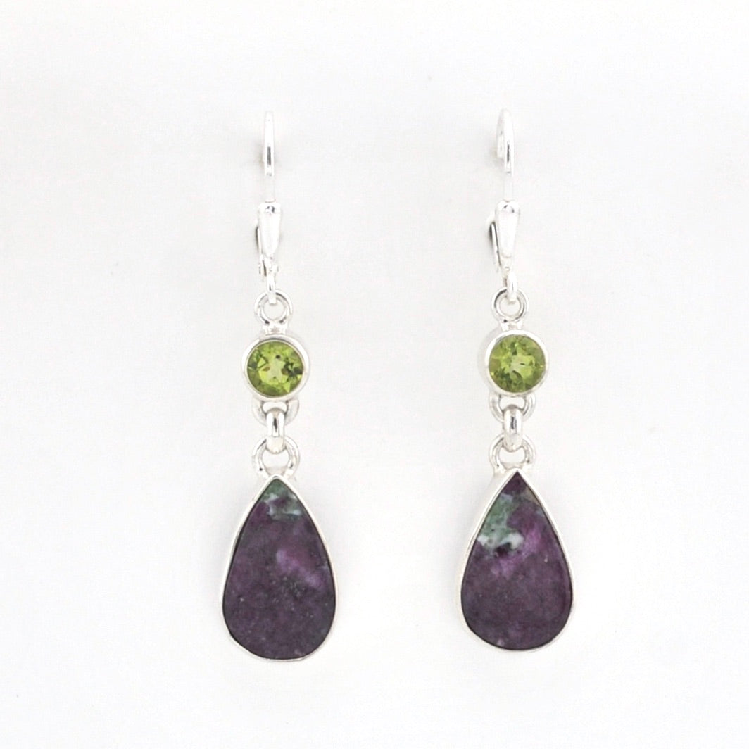 A pair of silver dangle earrings featuring a small round green gemstone and a larger teardrop-shaped dark purple stone, shown against a plain white background.