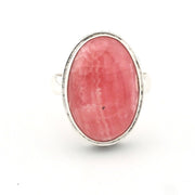 An oval ring featuring a pinkish-red cabochon gemstone in a silver bezel setting, shown against a plain white background.