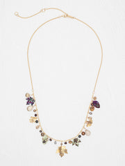 A gold-tone statement necklace featuring a delicate chain, a central cluster of mixed gemstones and charms in purple, green, brown, and clear, and two gold-tone leaf charms hanging at the bottom, displayed on a light textured background.