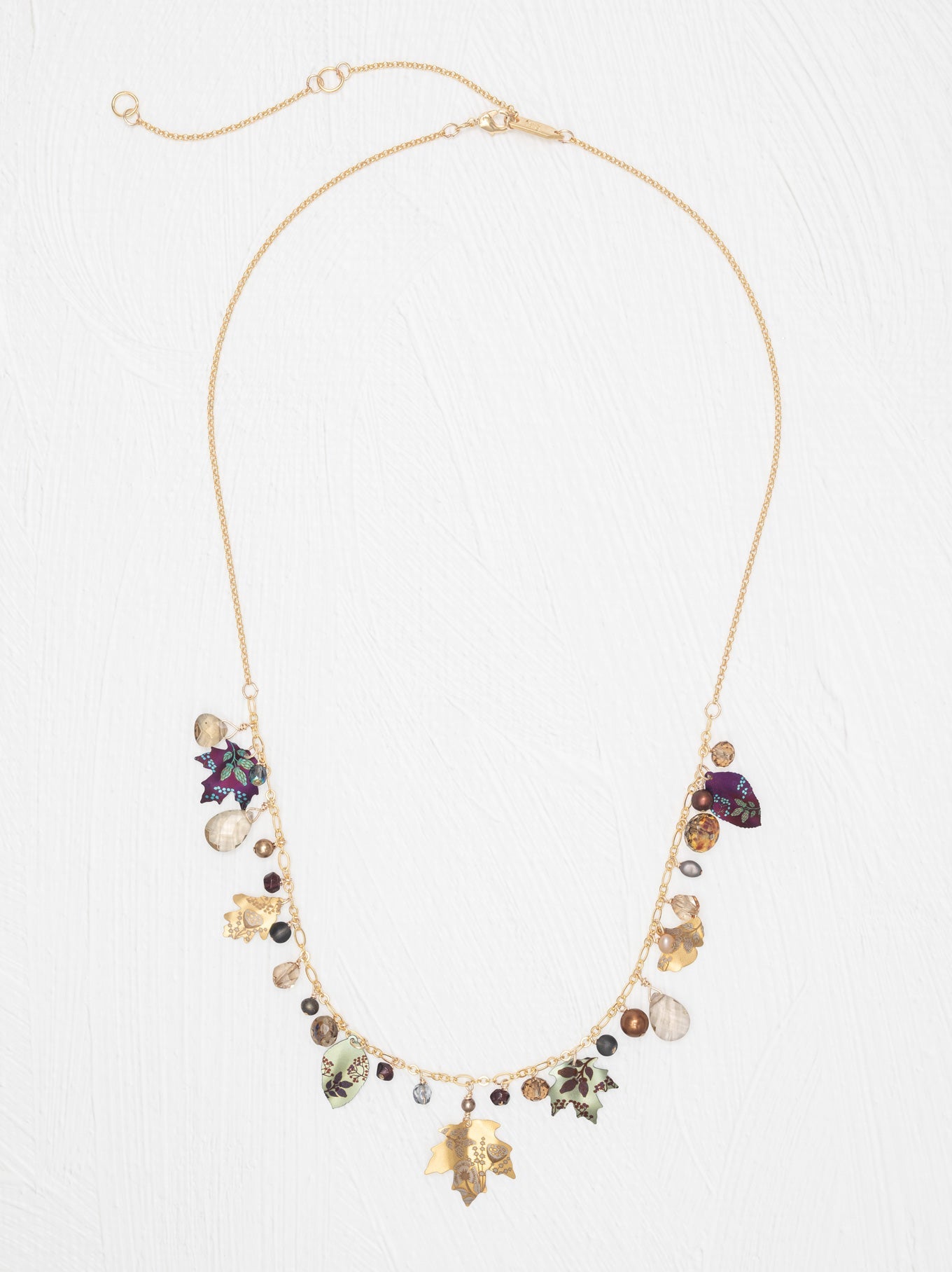 A gold-tone statement necklace featuring a delicate chain, a central cluster of mixed gemstones and charms in purple, green, brown, and clear, and two gold-tone leaf charms hanging at the bottom, displayed on a light textured background.