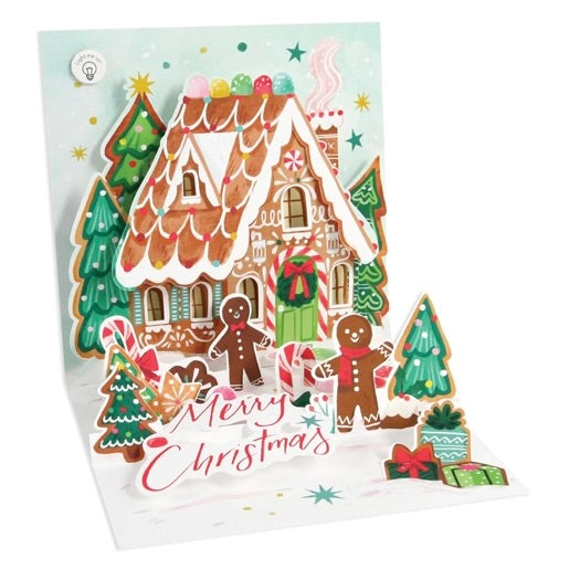 A 3D pop-up Christmas greeting card featuring a gingerbread house, candy canes, trees, and gingerbread people with the words 'Merry Christmas' written across the front.