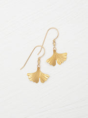 A pair of gold-tone dangle earrings with fan-shaped ginkgo leaf pendants, displayed on a light, textured white background.
