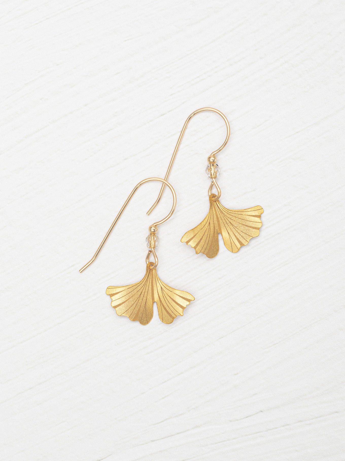 A pair of gold-tone dangle earrings with fan-shaped ginkgo leaf pendants, displayed on a light, textured white background.