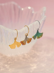 Three ginkgo leaf drop earrings displayed on a translucent white stand against a soft pink background. The earrings feature gold, rose gold, and green gradient leaf charms.