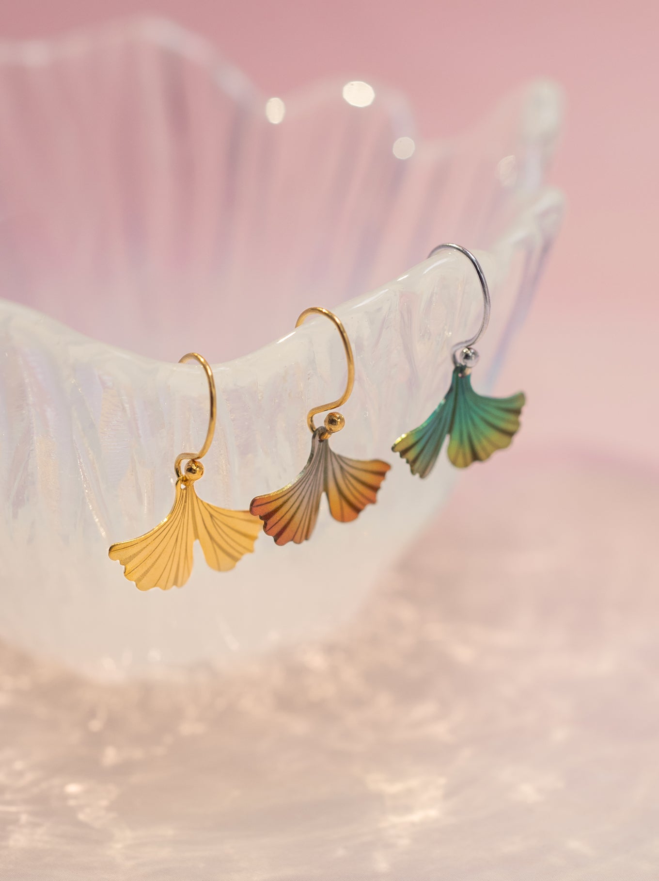 Three ginkgo leaf drop earrings displayed on a translucent white stand against a soft pink background. The earrings feature gold, rose gold, and green gradient leaf charms.