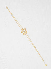 A delicate gold chain bracelet featuring a central gold flower charm with a clear faceted center, accented with small clear beads along the chain, laid out diagonally on a textured white background.