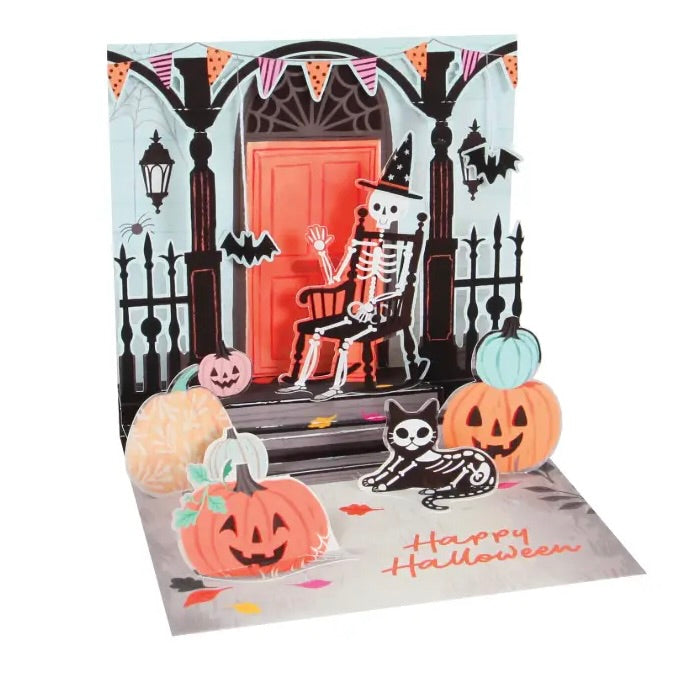 A 3D pop-up Halloween greeting card featuring a skeleton sitting on a porch, surrounded by jack-o'-lanterns, a black cat, bats, and the text 'Happy Halloween' on a white background.