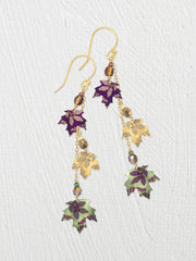 A pair of gold-tone dangle earrings with niobium-leaf charms in purple, gold, and sage green, accented with small faceted beads, shown on a textured white background.