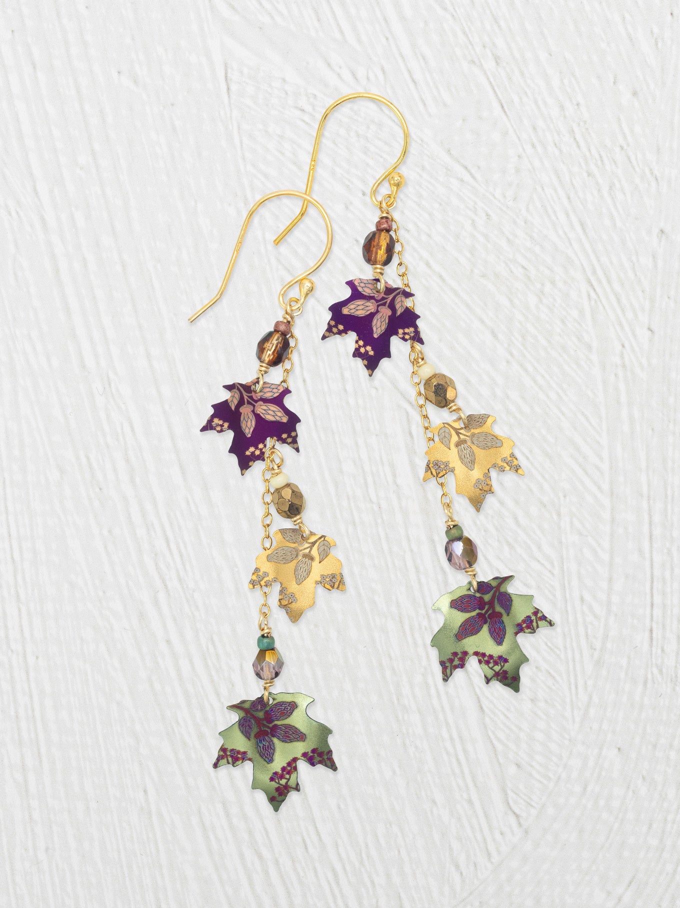 A pair of gold-tone dangle earrings with niobium-leaf charms in purple, gold, and sage green, accented with small faceted beads, shown on a textured white background.
