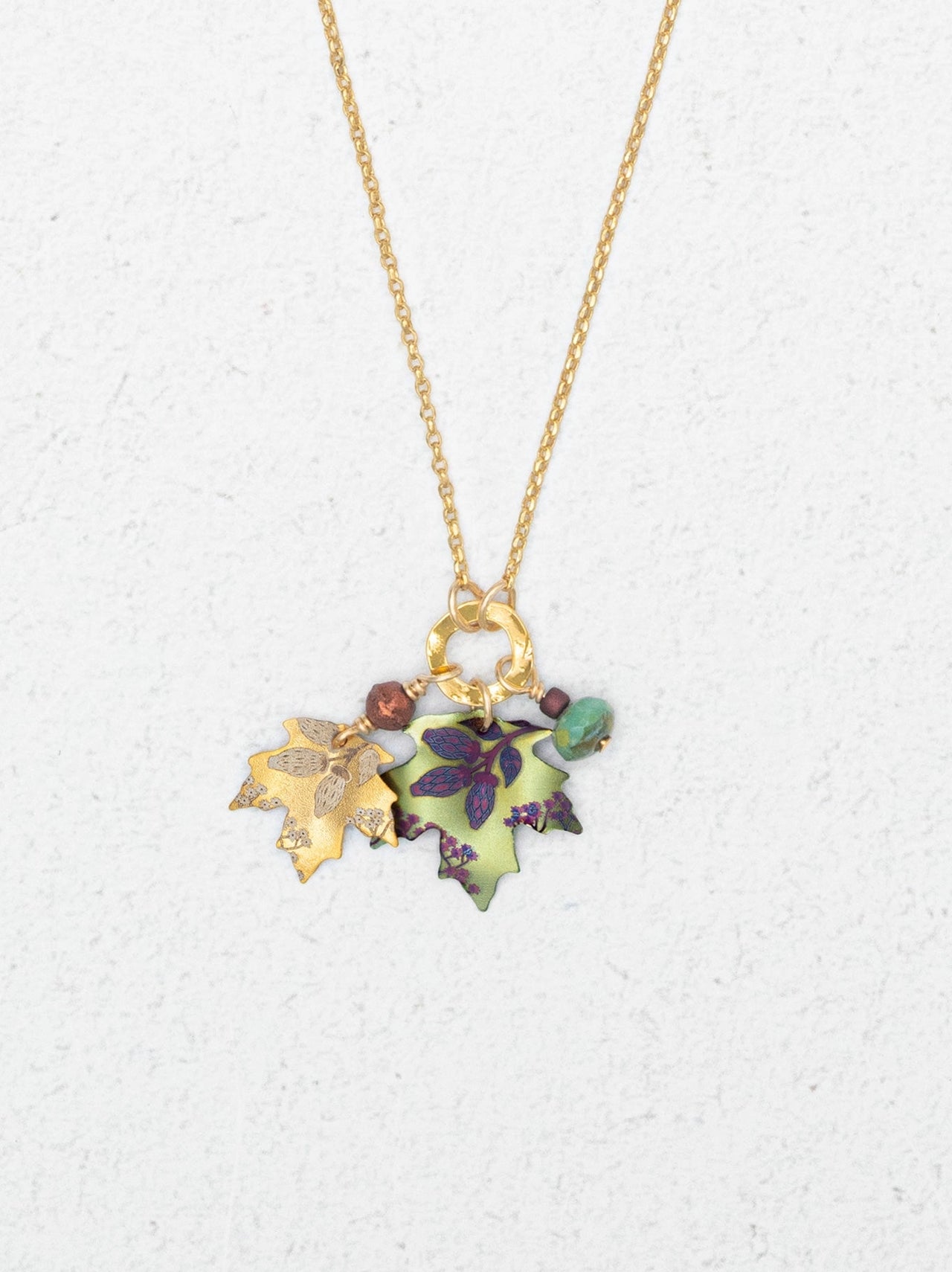 A gold chain necklace with three hanging niobium leaf charms in gold, green, and purple, accented with small beads, shown on a textured white background.