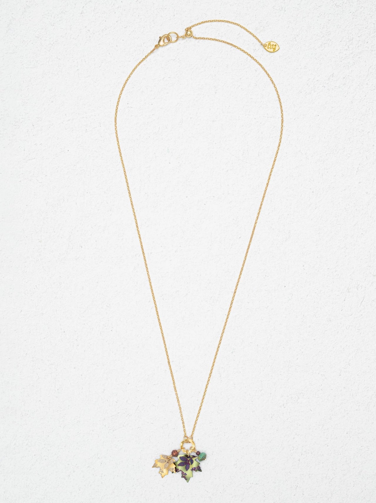A delicate gold chain necklace with a small floral cluster pendant featuring green, purple, and gold enamel details, displayed against a textured off-white background.