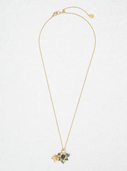 A delicate gold chain necklace with a small floral cluster pendant featuring green, purple, and gold enamel details, displayed against a textured off-white background.
