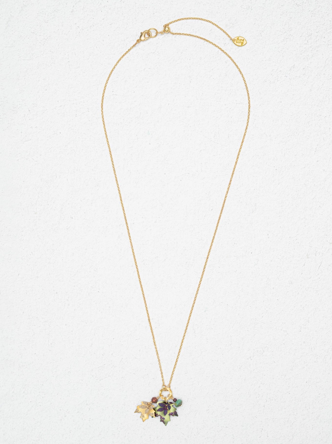 A delicate gold chain necklace with a small floral cluster pendant featuring green, purple, and gold enamel details, displayed against a textured off-white background.