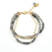 A multi-strand beaded bracelet featuring gray and blue-gray stone beads accented by gold-tone chain links, a lobster clasp, and a small dangling charm on a white background.