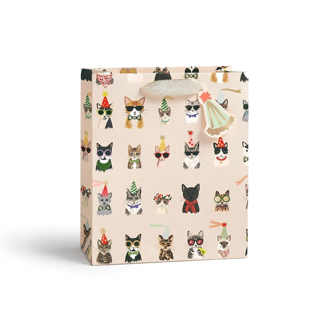 A small gift bag with a light beige background and an all-over print of illustrated cats in party hats and costumes. The bag has a silver glittery handle and is photographed on a clean white background.