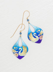 A pair of teardrop-shaped dangling earrings with gold-tone hooks and blue gradient panels featuring a white swan and gold floral details, displayed on a light wood background.