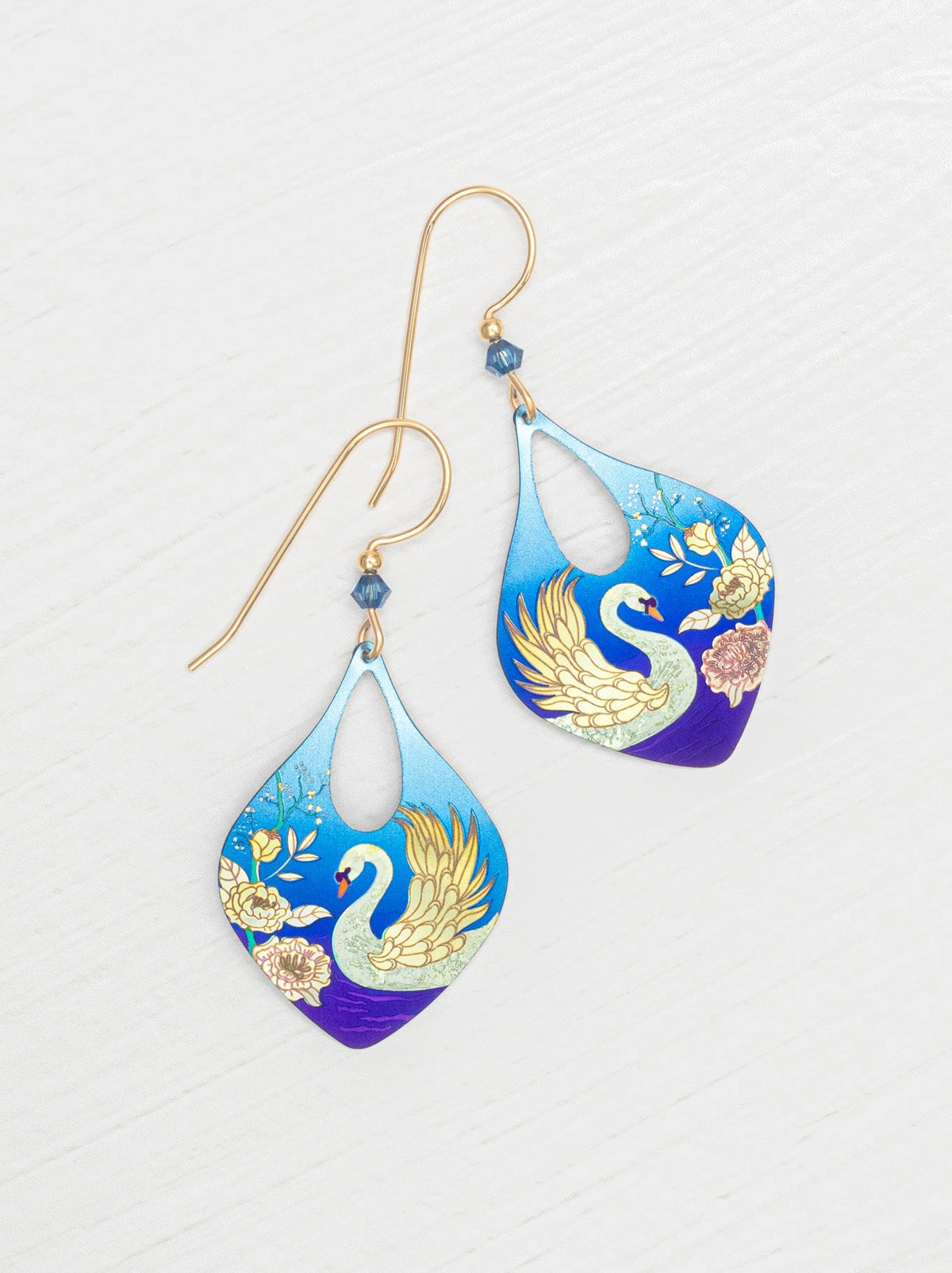 A pair of teardrop-shaped dangling earrings with gold-tone hooks and blue gradient panels featuring a white swan and gold floral details, displayed on a light wood background.