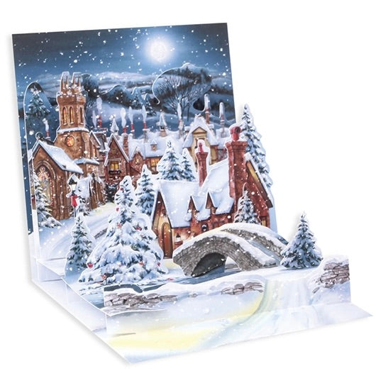 A 3D pop-up cardboard Christmas card featuring a snowy village scene with a church, cottages, snow-covered trees, a stone bridge, and a glowing full moon in the night sky.