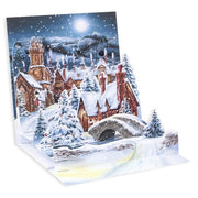 A 3D pop-up cardboard Christmas card featuring a snowy village scene with a church, cottages, snow-covered trees, a stone bridge, and a glowing full moon in the night sky.