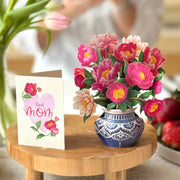 Vase of pink flowers on a wooden table with a card that says 'Best Mom' in a blurred indoor setting.