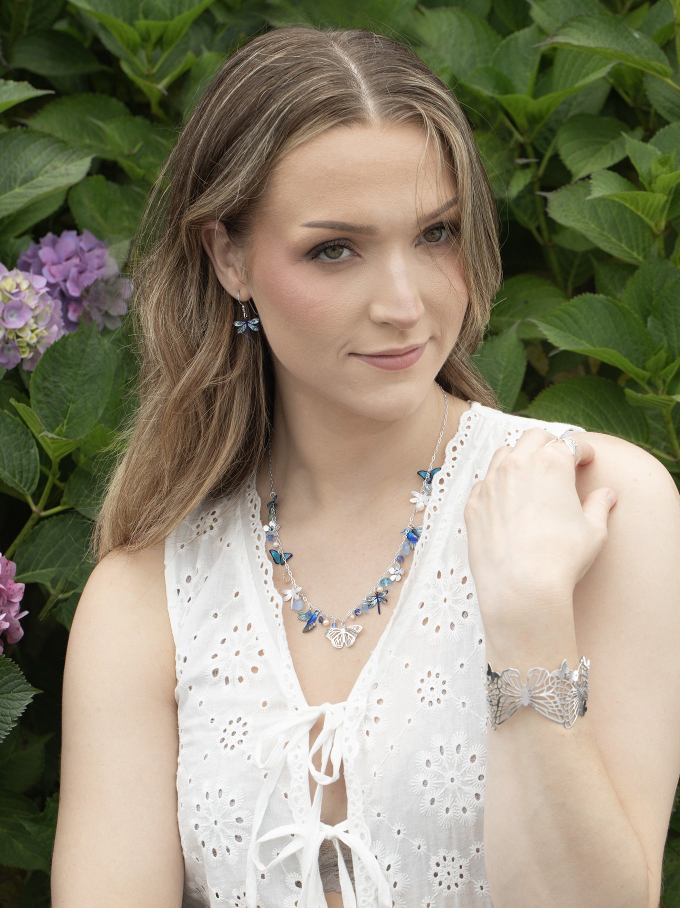 Woman wearing a white sleeveless top and jewelry against a green leafy background