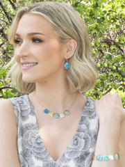 Woman with blonde hair wearing a floral dress and colorful jewelry outdoors.