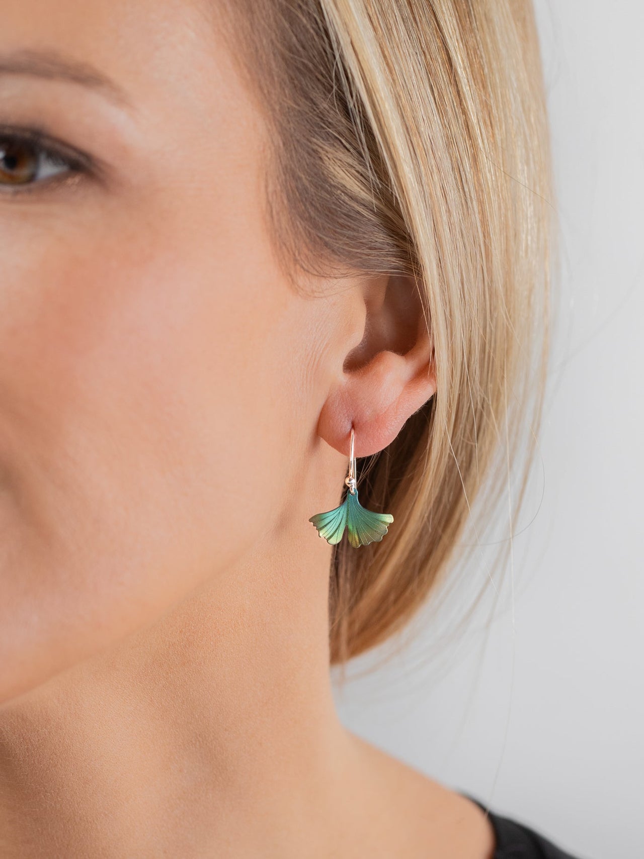 A close-up of a woman's ear wearing a small dangling earring with a green and blue fan-shaped charm on a gold hook, against a light gray background.